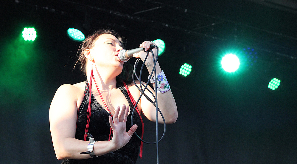 Image of Tanya Tagaq performing at Supercrawl 2017, singing into a microphone gripped in left hand along with a serpentine coil of mic cable, right arm bent at elbow, palm toward the audience. The artist wears a black dress and earrings with long red tassle ornamentation, a beaded cuff on left forearm and a silver cuff on right forearm featuring a pair of back-swept wolf claws. The background is dark, illuminated only by emerald green stage lighting.