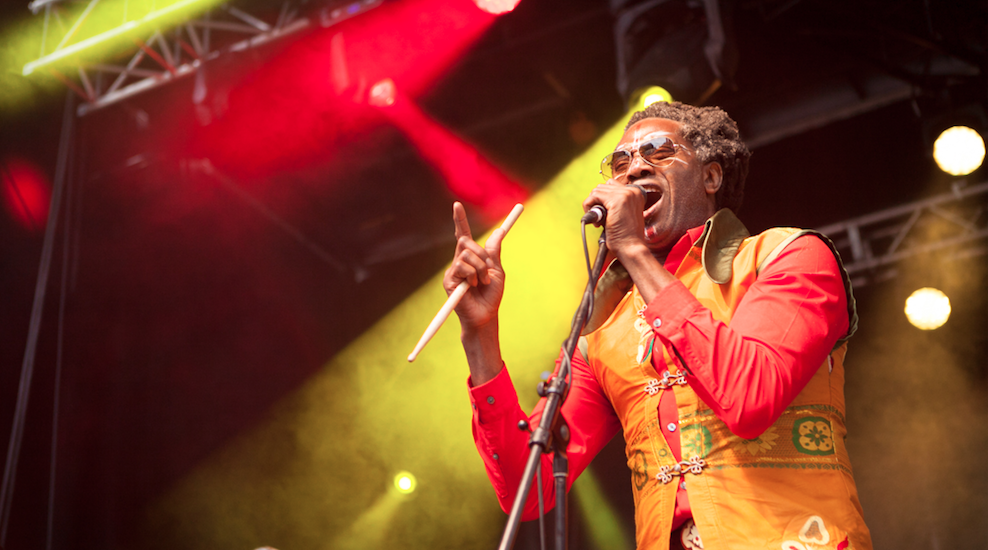 Image of Sifu Amayo, lead singer of Antibalas, performing at Supercrawl 2014. Stage lighting is a warm mix of reds and yellows. Amayo is wearing a red shirt and golden-orange vest, singing into a microphone that he holds in his left hand while holding a drum stick in his left hand, index finger pointed skyward. 
