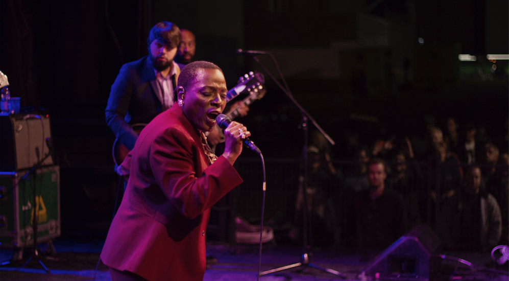 Image of Sharon Jones performing at Supercrawl 2015, with members of backing band The Dap-Kings and members of festival audience visible behind her.