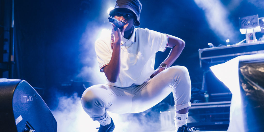 Image of artist Haviah Mighty of The Sorority at Supercrawl 2019. The singer squats in a low haze of wispy stage smoke, positioned between a pair of stage monitors. She gazes into the audience and raps into a wireless microphone held in her left hand, while her right hand is positioned on her waise. She is wearing a black bucket hat, loose-fitting white short-sleeve polo tee, pale-grey sweatpants cuffed above the ankle, white athletic socks and black sneakers. Stage lighting is cool white and gives the performer a glowing aura.