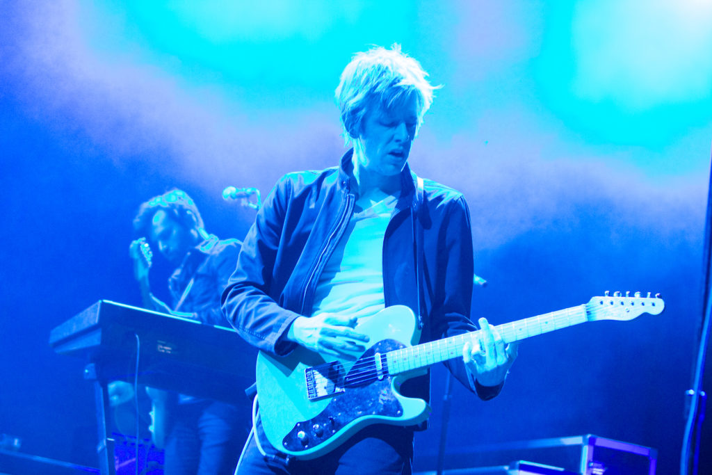 Image of Spoon performing at Supercrawl 2014, with lead singer Brit Daniel captured mid-riff, looking down toward the position of his left hand on the frets. Bassist Rob Pope is visible behind him, turned in the opposite direction. Stage lighting is a wash of cool blues. 