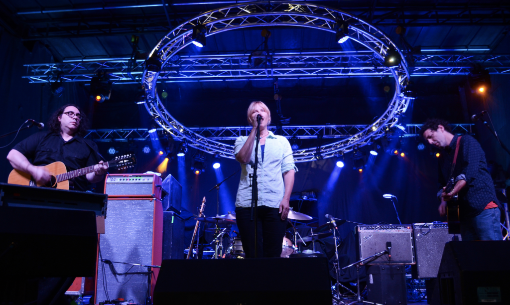 From left to right, James McNew, Georgia Hubley and Ira Kaplan of Yo La Tengo performing at Supercrawl 2013. Hubley stands at centre stage in a pale dress shirt with sleeves rolled above the elbow, singing into a microphone. A circular lighting truss is visible above and behind her like a halo. She is flanked by McNew and Kaplan in black shirts, who are playing acoustic and electric guitars respectively. 